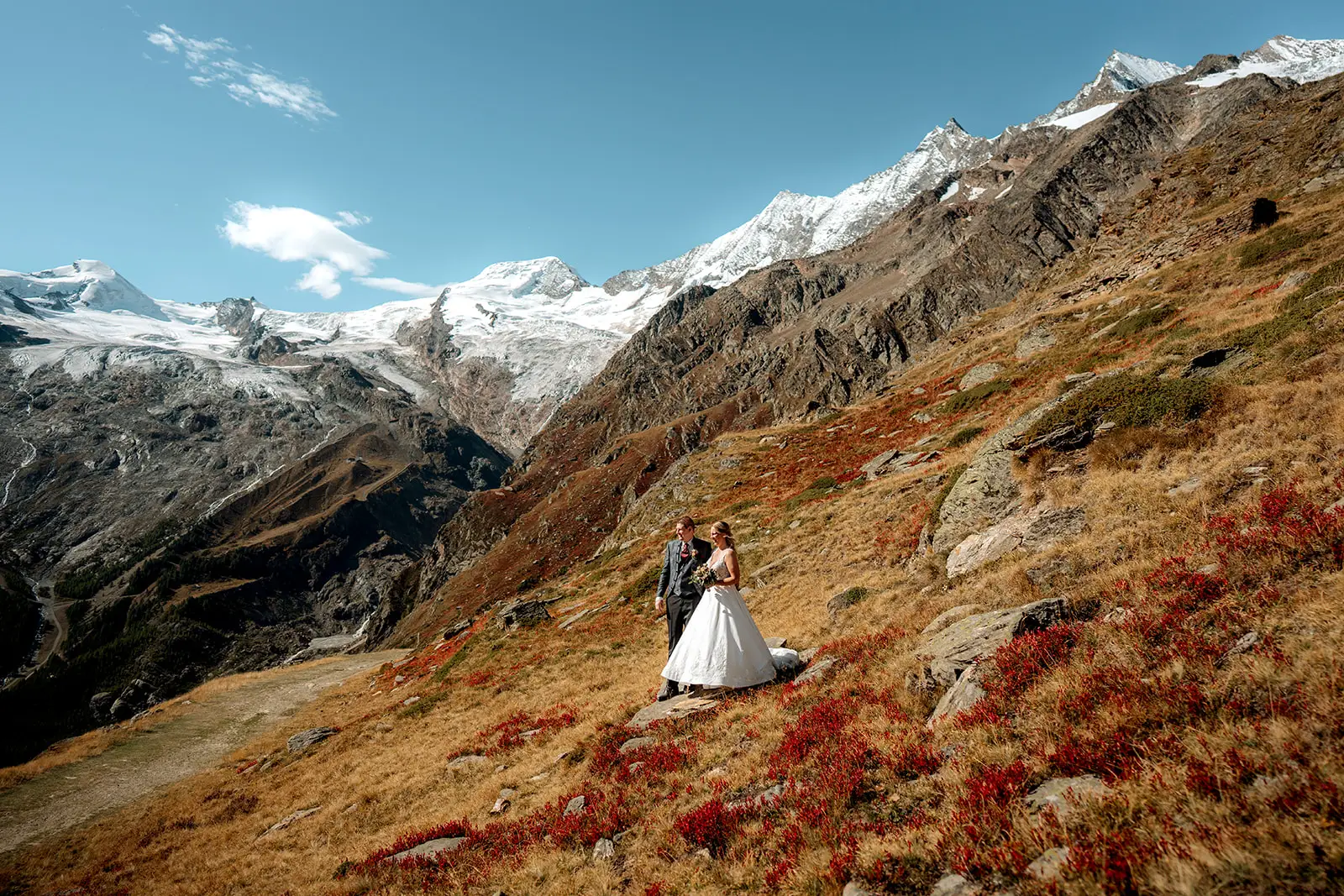 Eine Braut und ein Bräutigam stehen auf einem grasbewachsenen, felsigen Hügel mit schneebedeckten Bergen hinter ihnen, unter einem klaren blauen Himmel. Der hochzeitsfotograf fängt diesen atemberaubenden Moment ein, perfekt für jede hochzeitsreportage von denen, die sich entscheiden, in der Natur zu heiraten.