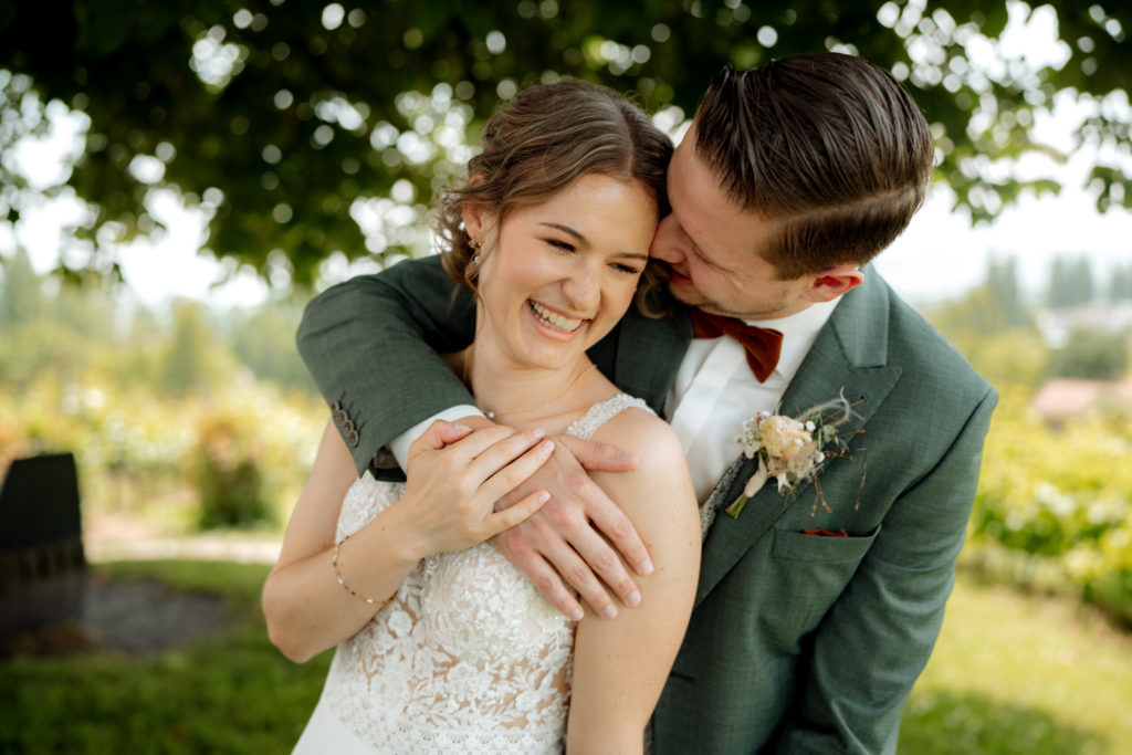 Eine freudige Umarmung zwischen Braut und Bräutigam im Freien. Aufgenommen von einem hochzeitsfotograf in Luzern, umarmt der Bräutigam die lächelnde Braut von hinten, beide in Hochzeitskleidung unter einem belaubten Baum mit Grünzeug und verschwommener Landschaft im Hintergrund.
