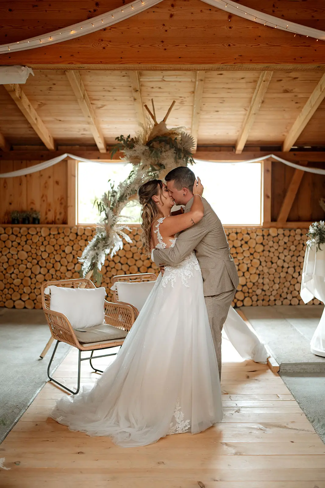 Eine Braut und ein Bräutigam küssen sich drinnen bei ihrer Hochzeit auf einem Holzboden vor einer rustikalen Kulisse aus gestapeltem Brennholz und Pampasgras, während natürliches Licht durch die Fenster fällt - perfekt für authentische Hochzeitsfotografie.