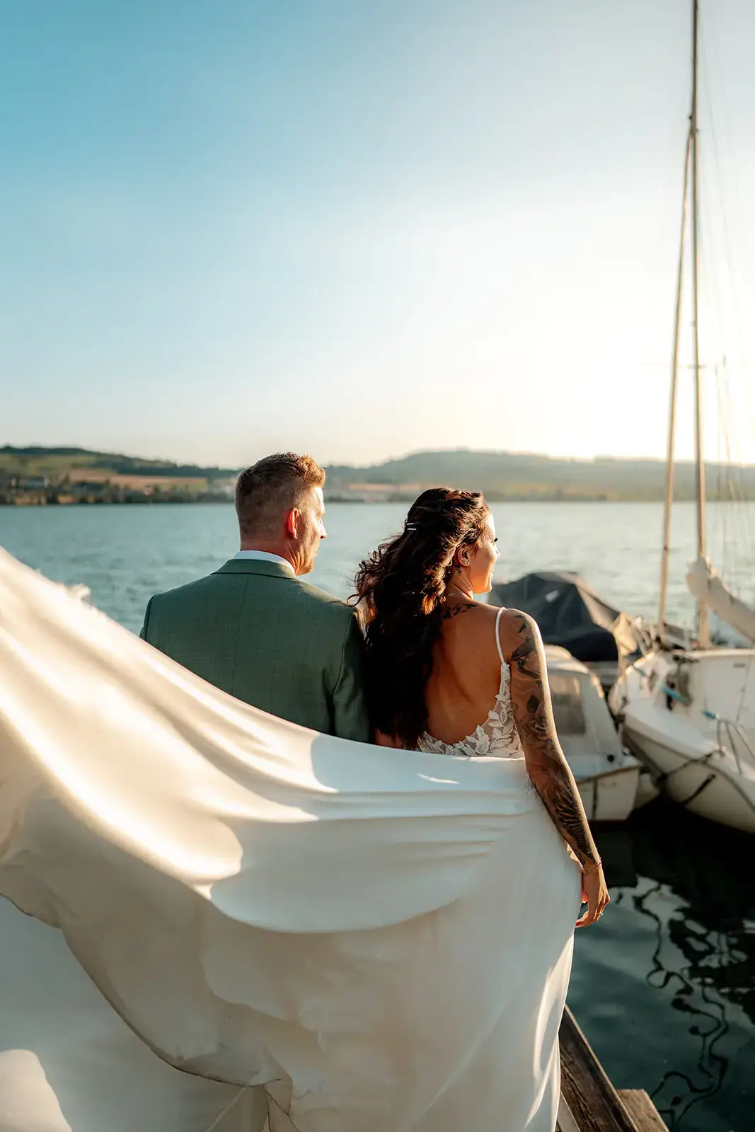 Eine Braut und ein Bräutigam stehen auf einem Steg mit Segelbooten in Luzern und blicken aufs Wasser. Das fließende weiße Kleid der Braut wird von der Brise erfasst, während die Sonne über dem See untergeht - perfekt für unvergessliche Hochzeitsfotografie und diejenigen, die vom Heiraten träumen.