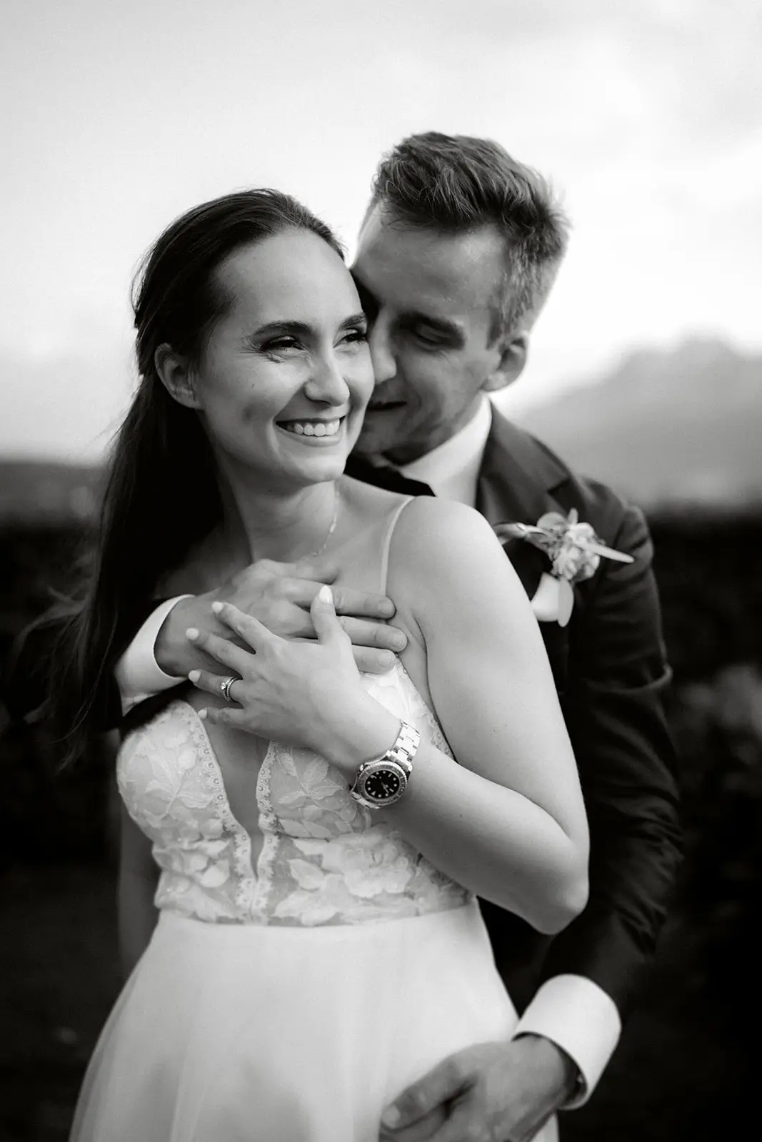 Eine Braut und ein Bräutigam umarmen sich glücklich lächelnd im Freien. Eingefangen von einem hochzeitsfotograf, ist ihr freudiger Moment Teil einer zeitlosen hochzeitsreportage. Der Bräutigam steht hinter der Braut in diesem schönen schwarz-weiß Bild.