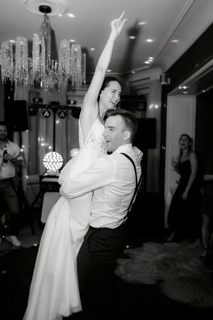 Ein Bräutigam hebt eine freudige Braut in einem weißen Kleid hoch, die feierlich einen Arm hebt. Sie tanzen auf ihrer Hochzeit mit Gästen und Kronleuchtern im Hintergrund - perfekt für jeden Hochzeitsfotografen, um sie festzuhalten.