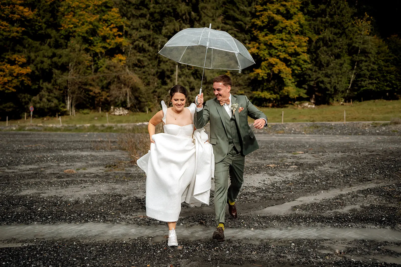 Eine Braut in einem weissen Kleid und Turnschuhen und ein Bräutigam in einem grünen Anzug laufen fröhlich unter einem durchsichtigen Regenschirm auf einem felsigen Weg, umgeben von Bäumen mit grünen und gelben Blättern, um ihre Hochzeit in Luzern zu feiern.