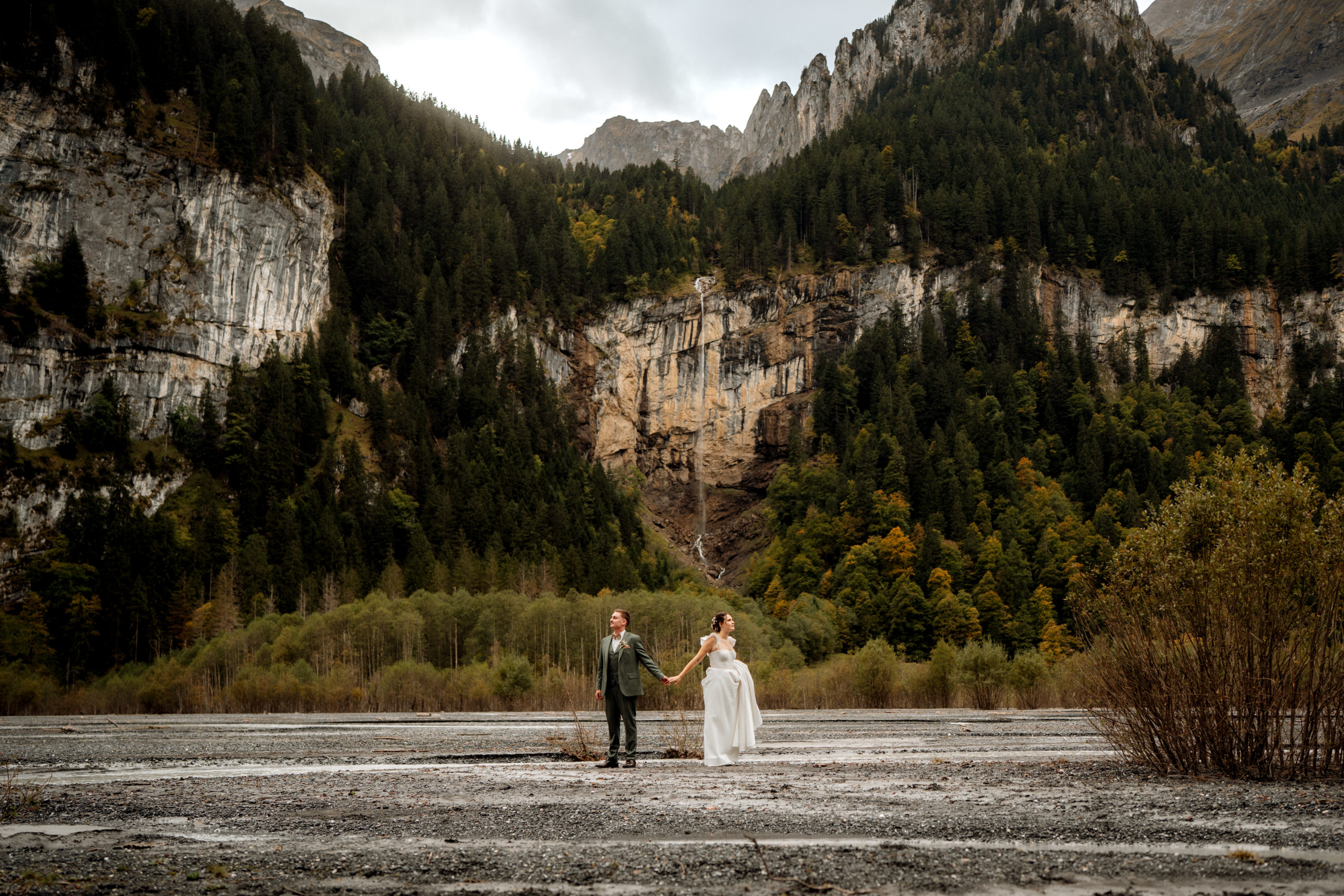 Eine Braut und ein Bräutigam stehen händchenhaltend im Vordergrund einer dramatischen Berglandschaft, perfekt für Hochzeitsfotografie, mit dichten grünen Wäldern, felsigen Klippen und bedecktem Himmel.