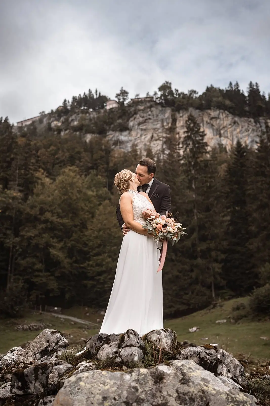 Eine Braut und ein Bräutigam stehen auf einem Felsen und umarmen sich, wobei die Braut einen Blumenstrauß hält. Umgeben von hohen Kiefern und Bergen unter einem wolkenverhangenen Himmel fängt diese Hochzeitsfotografie den Zauber des Heiraten in der Natur ein.