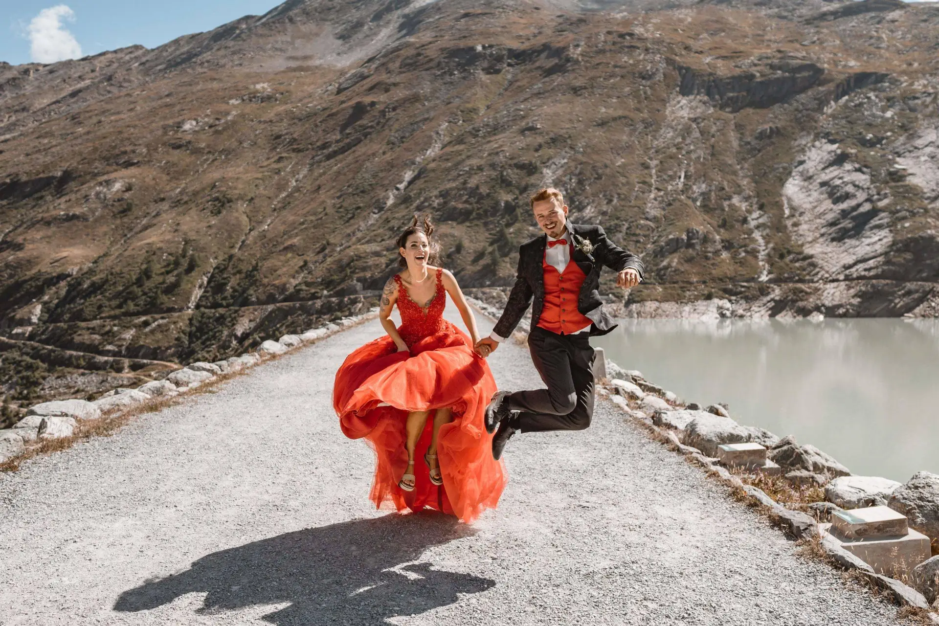 Eine Frau in einem roten Kleid und ein Mann in einem schwarzen Anzug mit roter Fliege hüpfen fröhlich auf einem Bergpfad neben einem See - und fangen den lebendigen Geist der Hochzeitsfotografie vor dem Hintergrund atemberaubender felsiger Hügel und eines klaren Himmels ein.