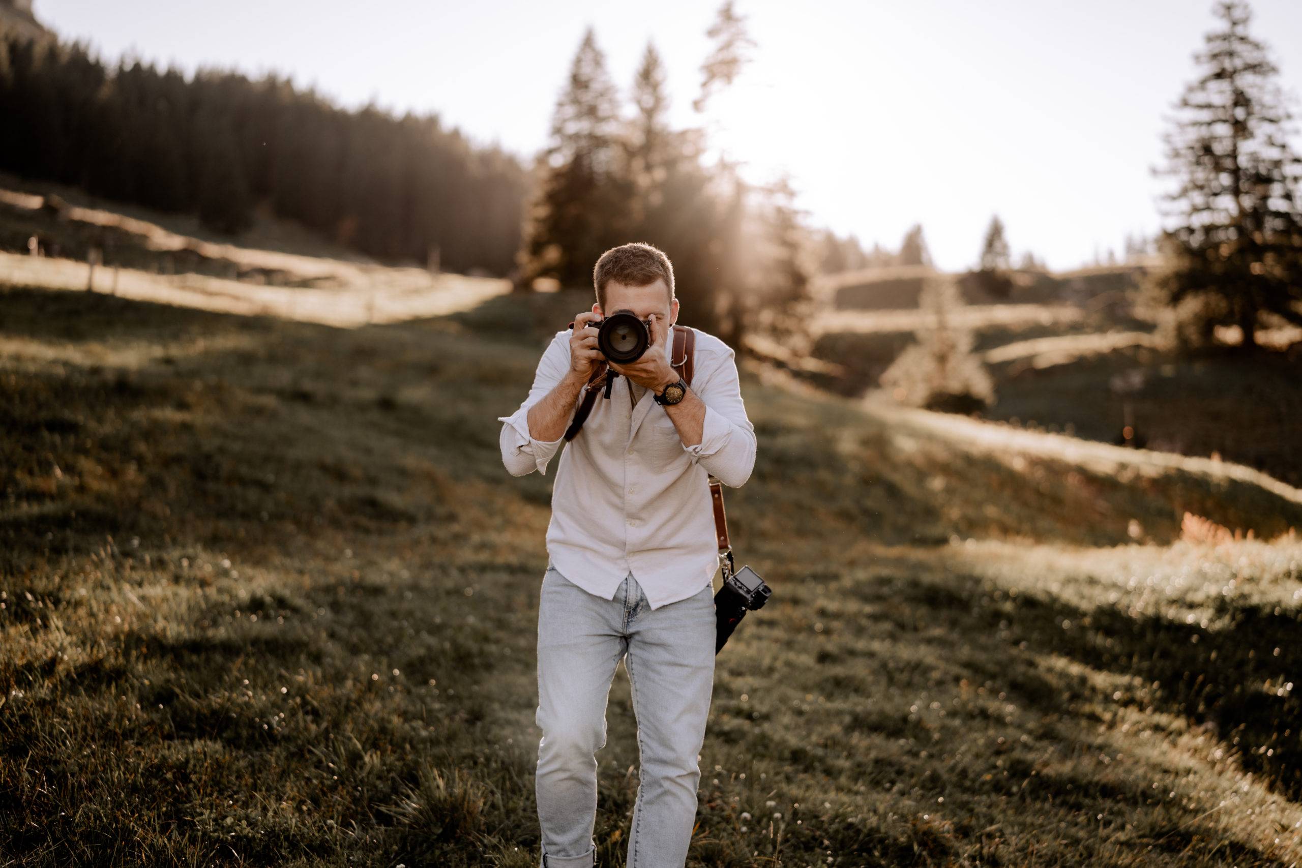 Eine Person in weißem Hemd und hellen Jeans steht auf einem grasbewachsenen Gelände und hält eine Kamera an ihr Gesicht - vielleicht ein Hochzeitsfotograf, der Erinnerungen festhält. Kiefern und Sonnenlicht füllen den Hintergrund und schaffen eine warme Atmosphäre im Freien.