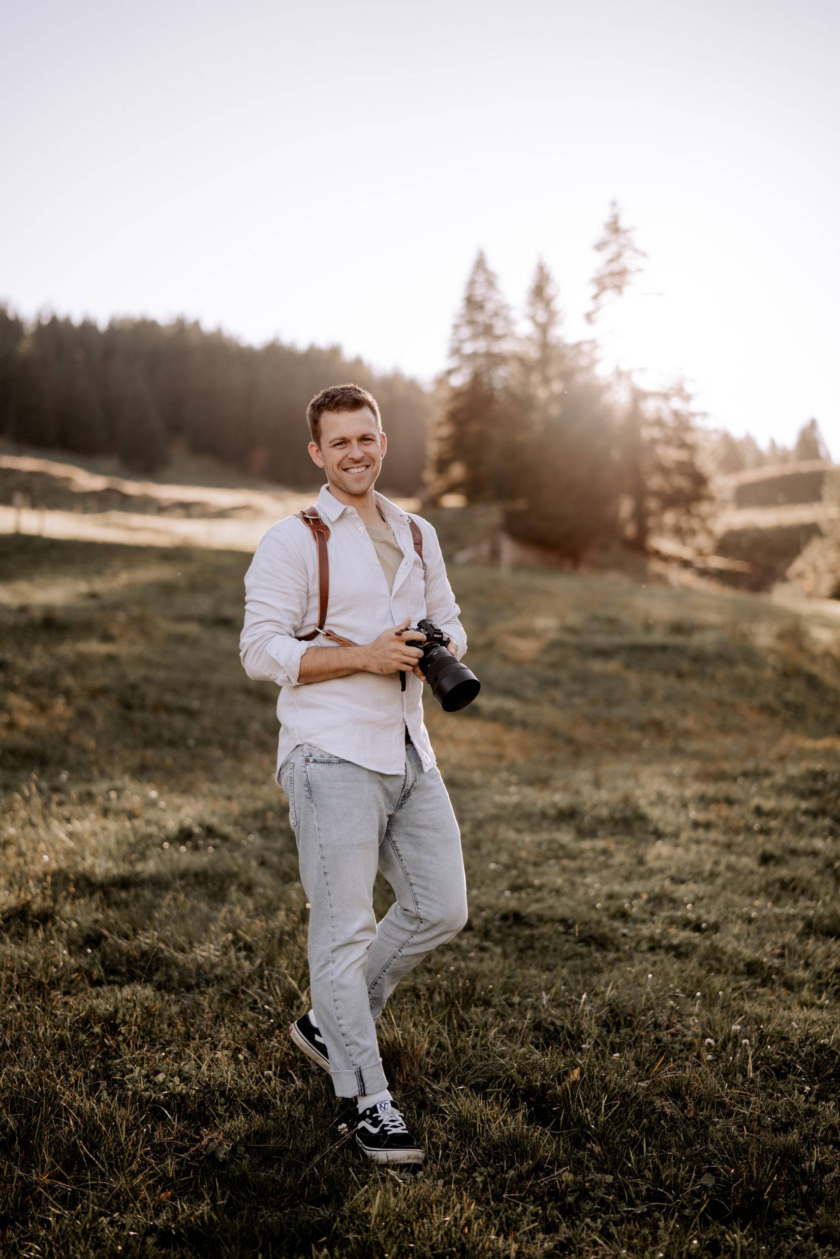 Ein Mann in weißem Hemd und hellen Jeans steht mit der Kamera in der Hand auf einer Wiese in Luzern. Im Hintergrund fällt das Sonnenlicht durch die Bäume - perfekt, um unvergessliche Hochzeitsreportage-Momente festzuhalten.