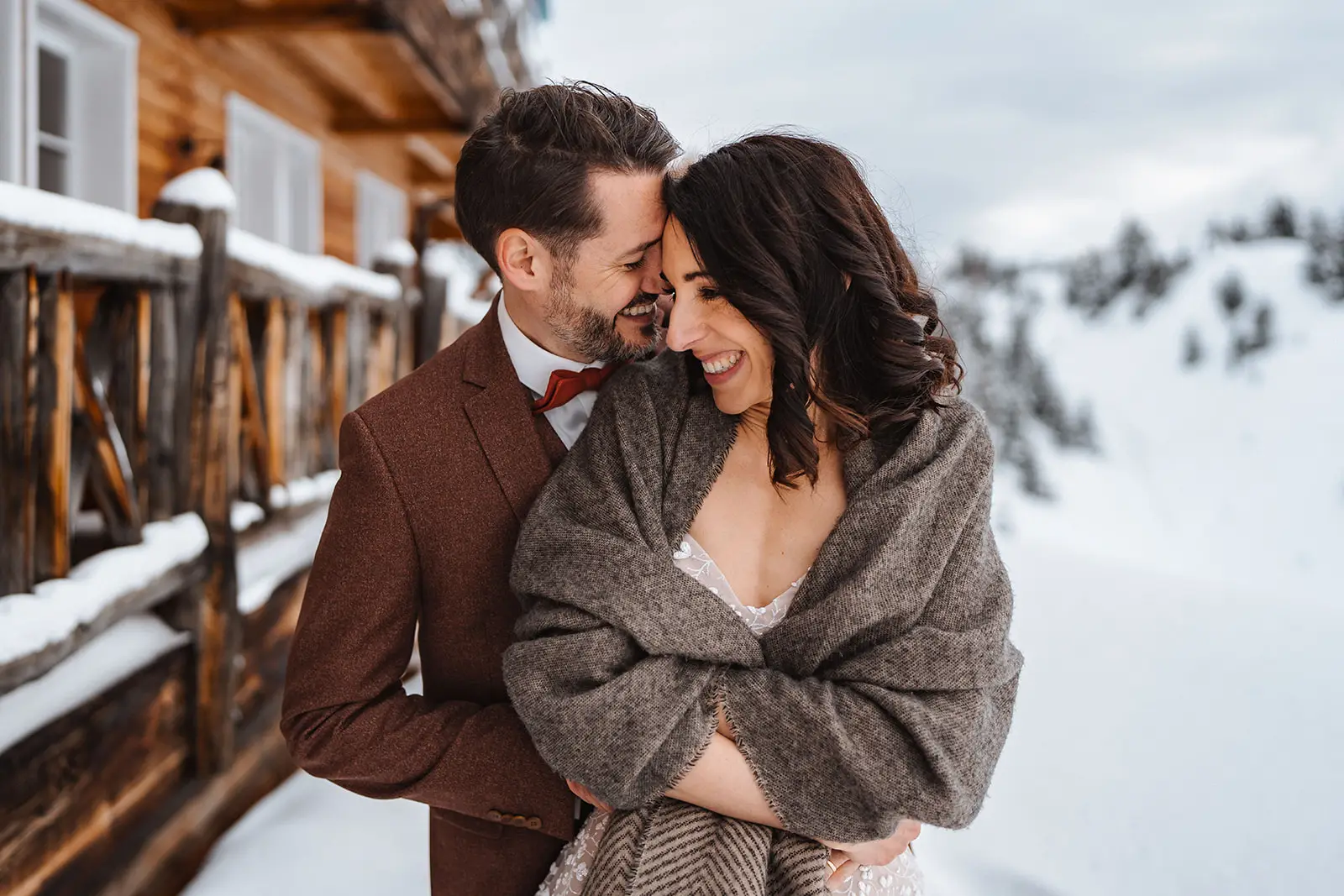 Ein lächelndes Paar umarmt sich im Freien in einer verschneiten Berglandschaft bei Luzern. Der Mann im braunen Anzug hält die in einen grauen Schal gehüllte Frau, während sie vor einer Holzhütte stehen und die Wärme ihrer Liebe in diesem hochzeitsreportage-Moment festhalten.