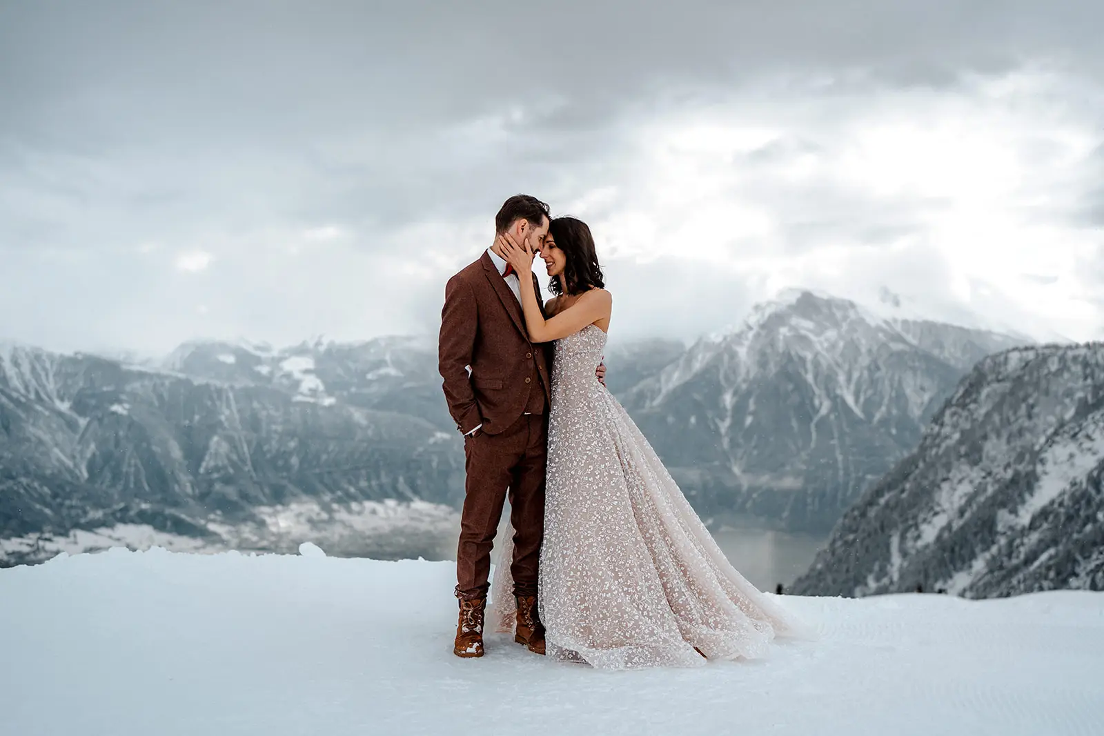 Ein Paar steht eng beieinander auf einem verschneiten Berg, die malerischen Gipfel im Rücken, während ein Hochzeitsfotograf den Moment festhält. Das elegante Kleid der Frau und der braune Anzug des Mannes spiegeln den Zauber von heiraten in dieser atemberaubenden Winterlandschaft wider.