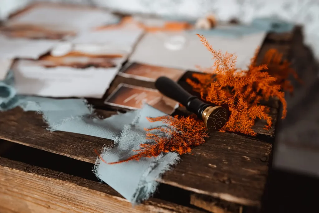 Auf einem Holztisch liegen zerrissener blauer Stoff, orangefarbenes getrocknetes Laub, alte Briefumschläge, Karten und ein Wachssiegelstempel aus Messing - die perfekte Inspiration für eine rustikale Hochzeit oder zum Festhalten mit Ihrem Hochzeitsfotografen.