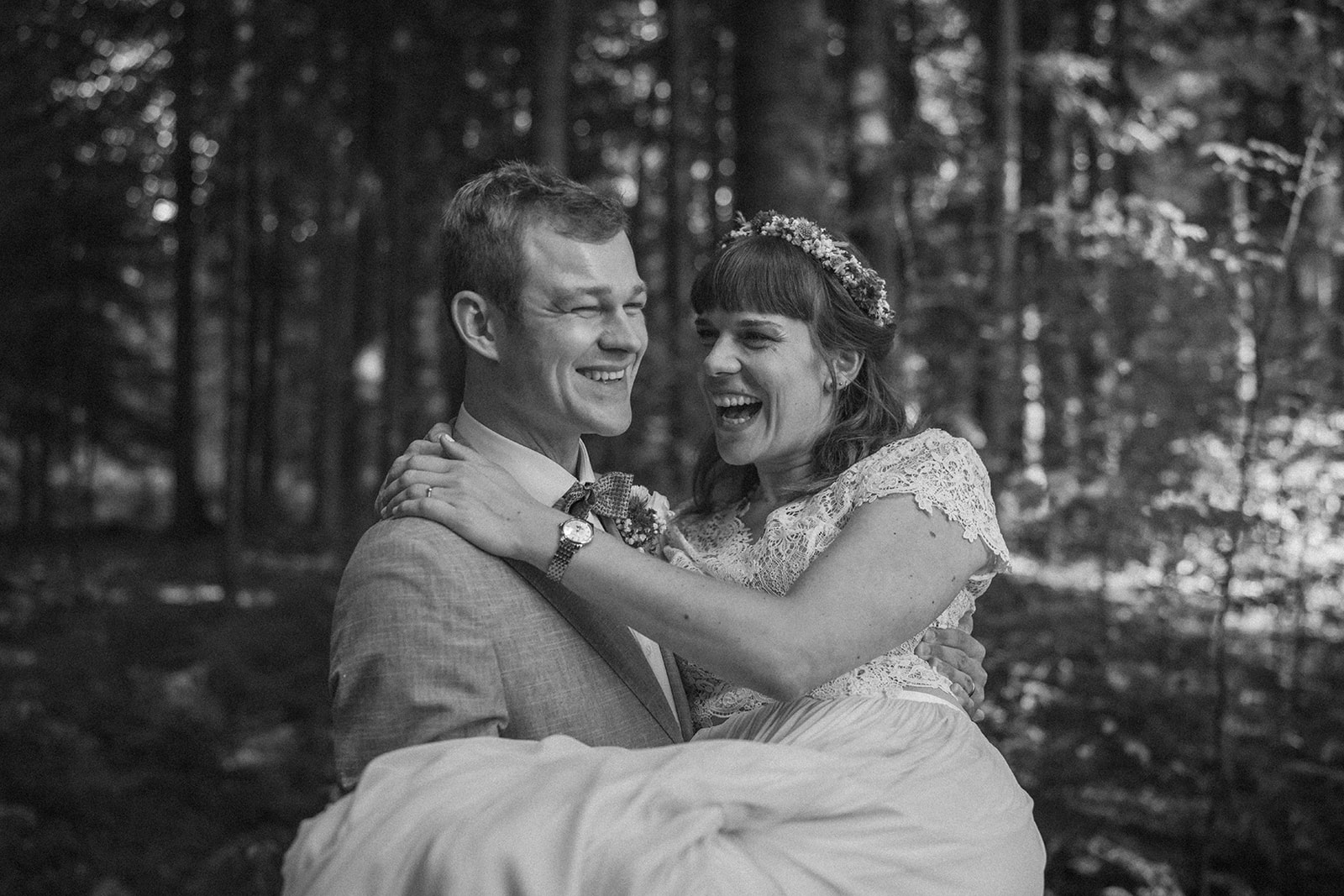 Ein Bräutigam in einem hellen Anzug lächelt, während er eine lachende Braut in einem Spitzenhochzeitskleid und einer Blumenkrone vor einem Waldhintergrund in weichem, natürlichem Licht trägt.