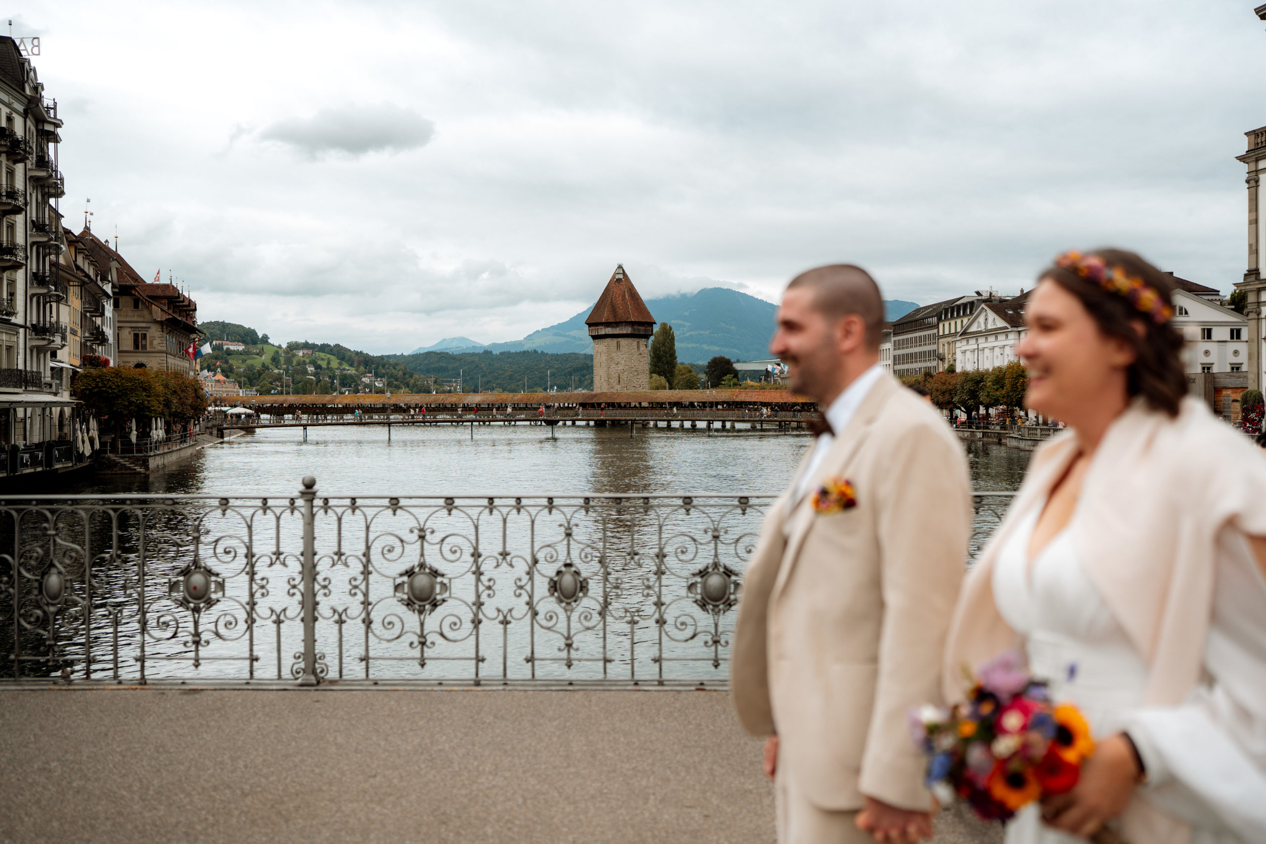Ein Paar in Hochzeitskleidung steht auf einer Brücke an einem malerischen Fluss, mit historischen Gebäuden, einem Turm und Bergen im Hintergrund unter einem bewölkten Himmel. Der Fokus liegt auf dem Hintergrund, während das Paar im Vordergrund verschwommen ist.