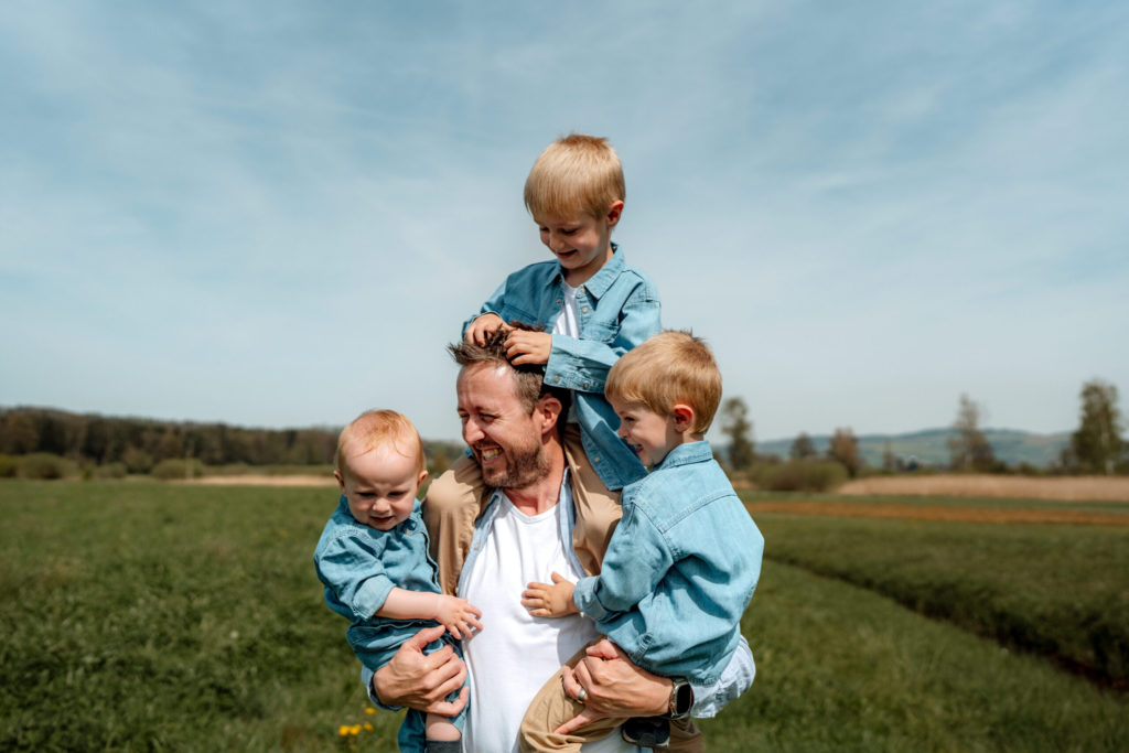 Ein lächelnder Mann steht auf einer grünen Wiese und hält zwei kleine Kinder in Jeanshemden, während ein drittes Kind, ebenfalls in Jeans, auf seinen Schultern sitzt und an einem sonnigen Tag mit seinem Haar spielt.