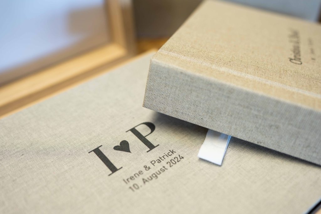 Nahaufnahme eines beigen Fotoalbums mit I ♥ P und den Namen Irene & Patrick sowie dem Datum 10. August 2024 aufgedruckt. Ein weiteres Album mit einem weißen Band ist teilweise oben sichtbar.