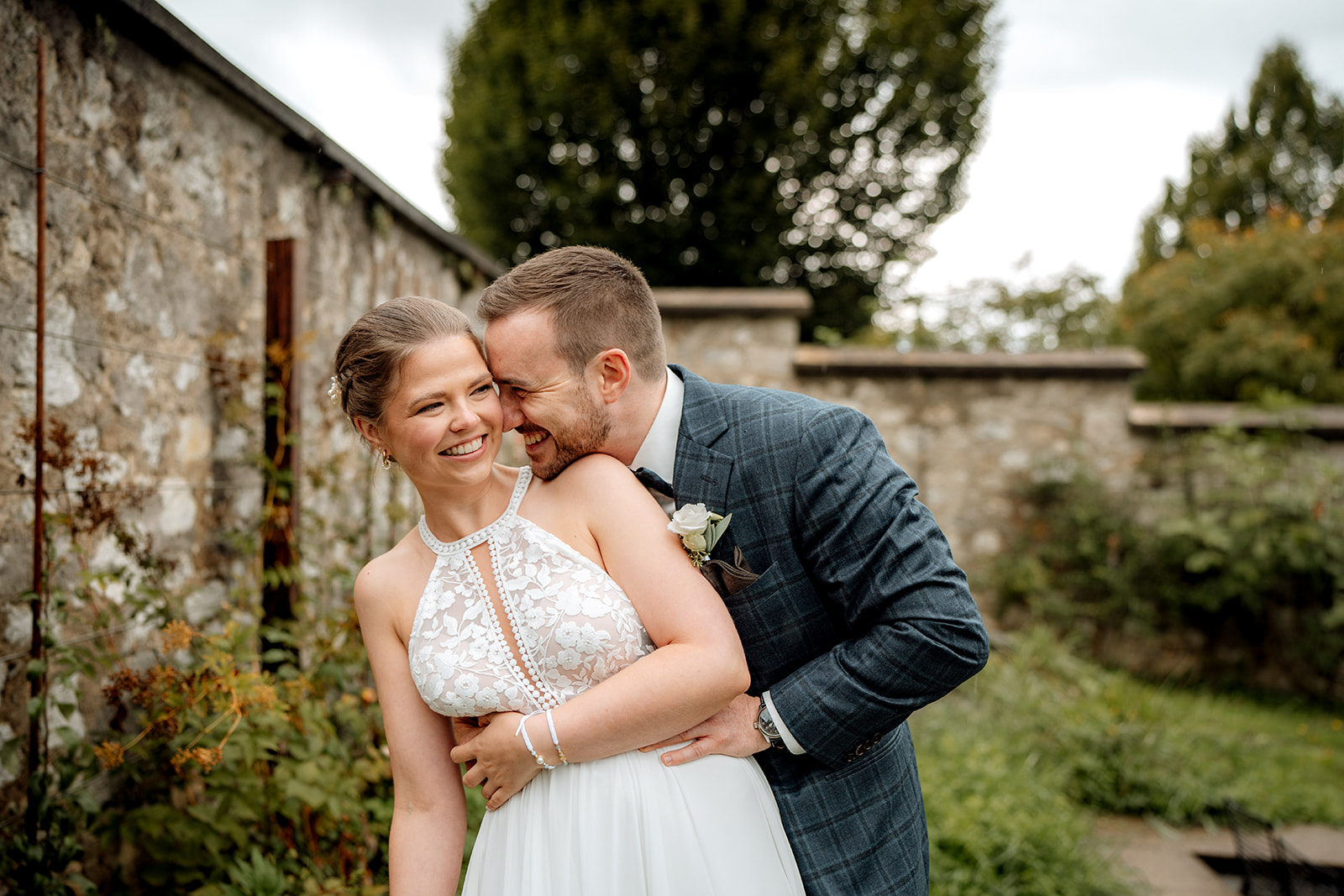 Eine lächelnde Braut in einem weißen Spitzenkleid steht im Freien, während der Bräutigam, der einen blaukarierten Anzug trägt, sie von hinten umarmt und sich spielerisch an sie anlehnt. Sie wirken fröhlich in einem Garten mit Steinmauern und Grünpflanzen.