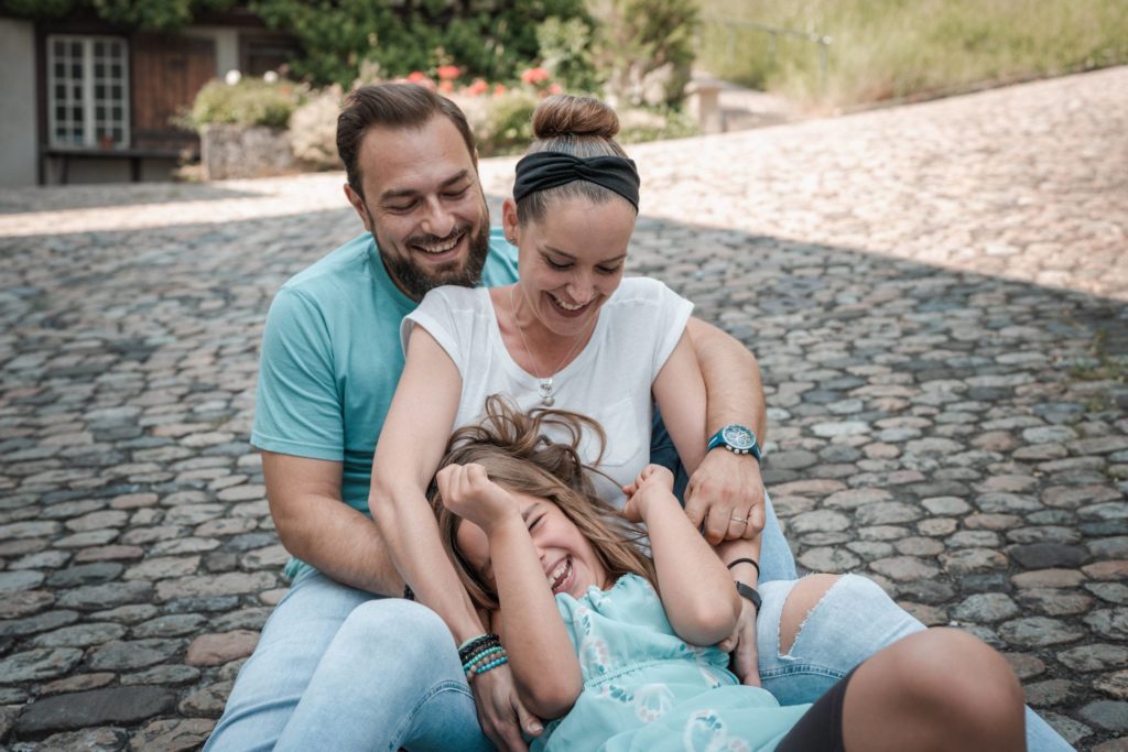 Ein lächelnder Mann und eine lächelnde Frau sitzen auf einem Kopfsteinpflasterweg und kitzeln und umarmen spielerisch ein lachendes junges Mädchen vor ihnen. Alle drei genießen einen glücklichen Moment zusammen im Freien.